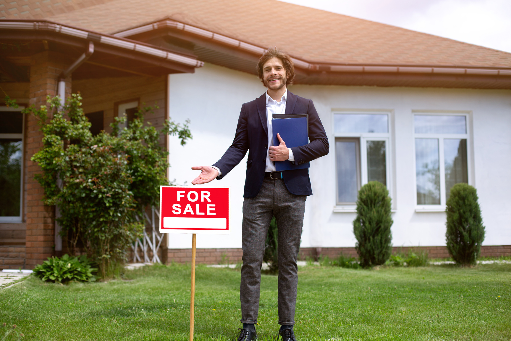 Cheerful property manager showing FOR SALE sign near country house